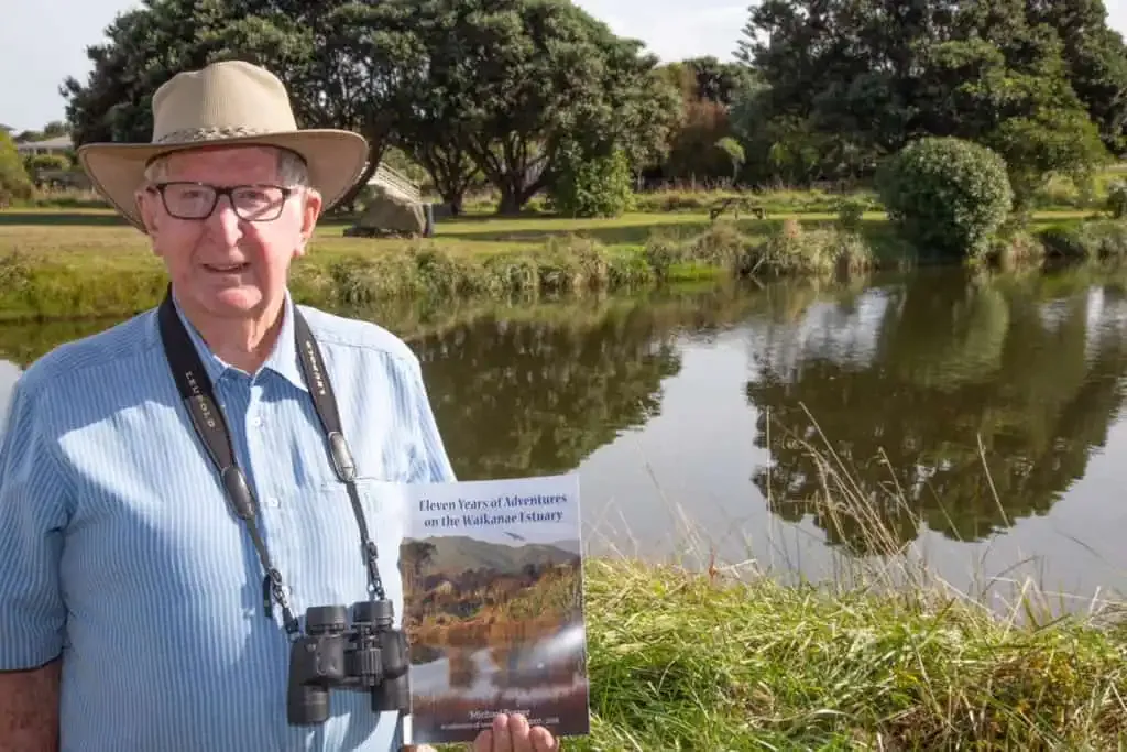 Mik Peryer with binoculars, holding one of his bird books beside the estuary